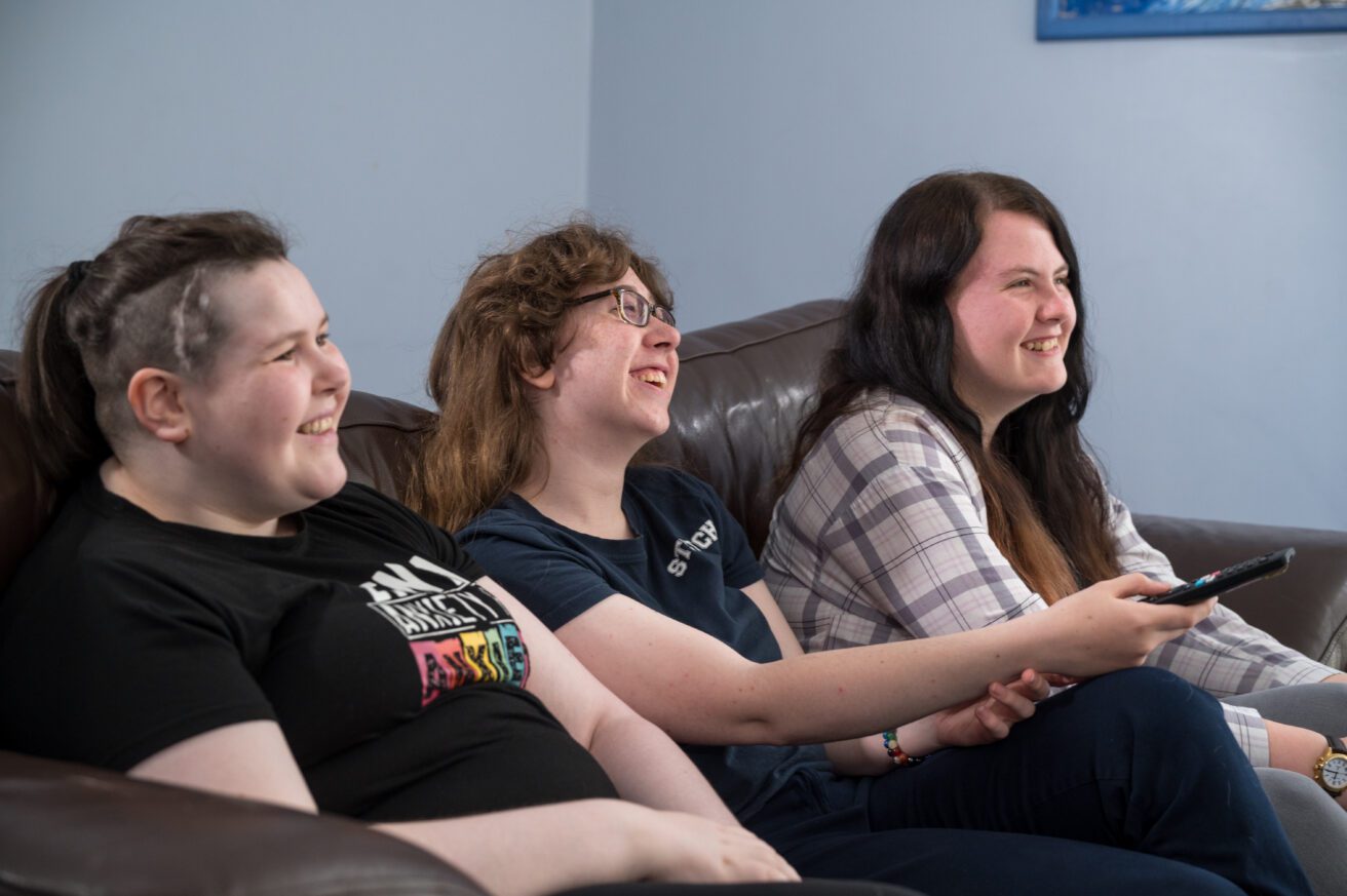 3 female students sitting on a sofa in residential halls. One is holding the TV remote out and pointing towards the TV