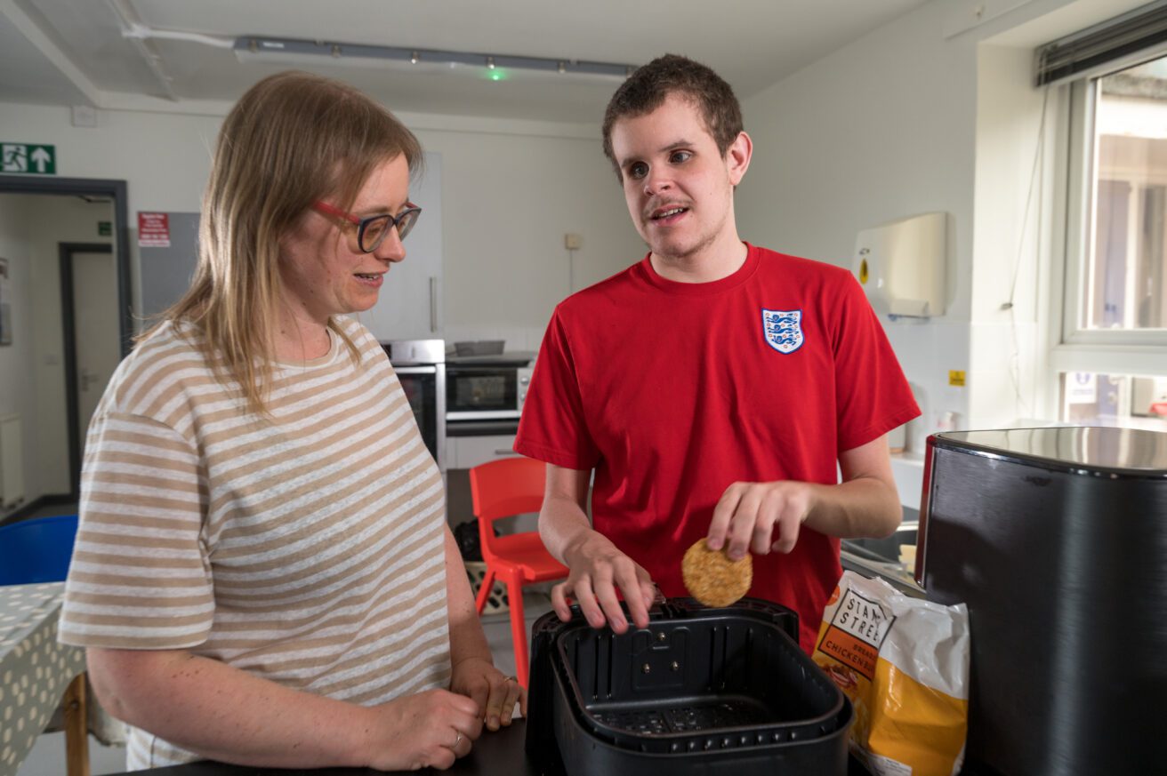 A student in a kitchen setting places a food item into the basket of an air fryer as his teacher watches on