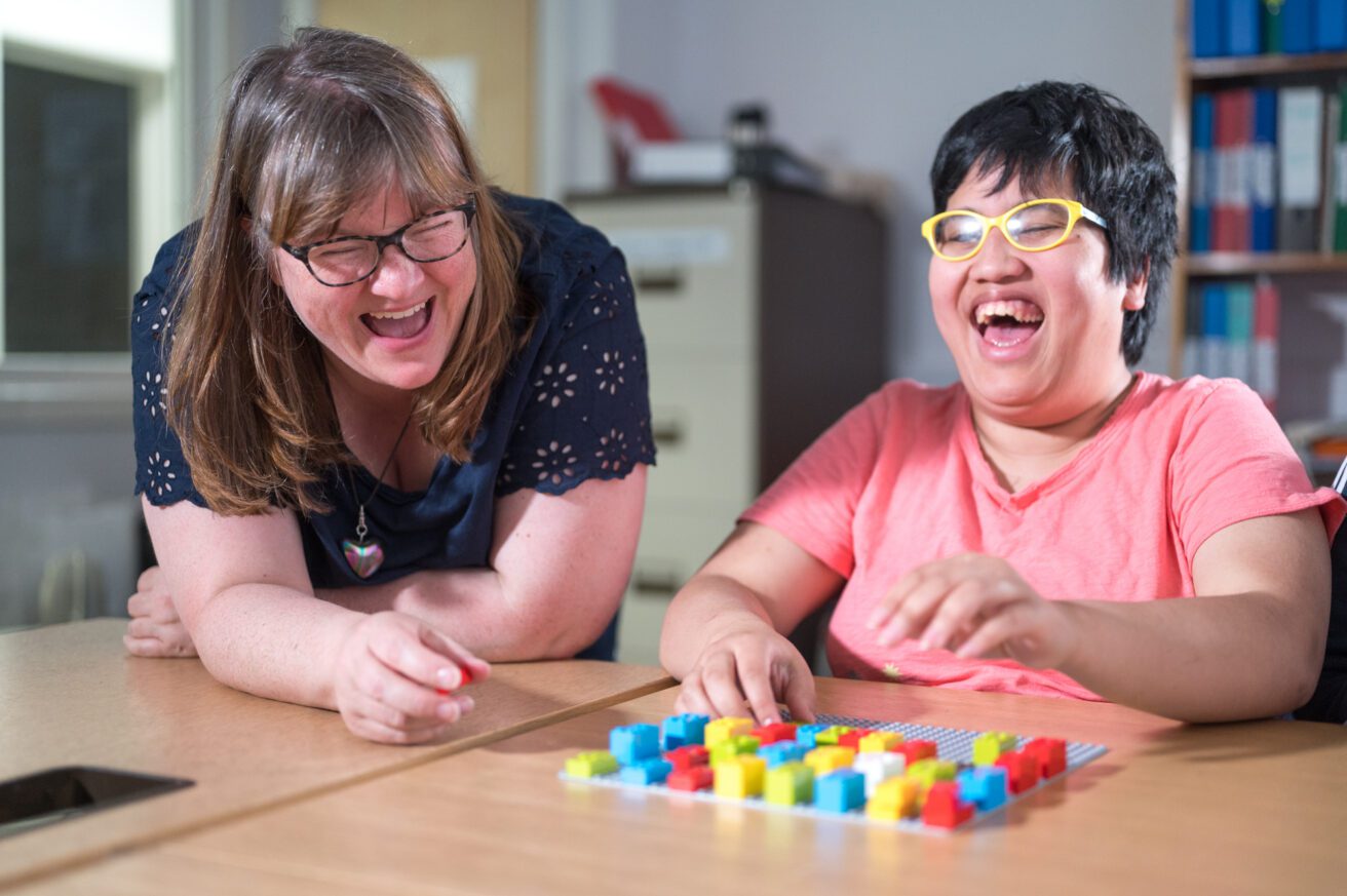 a female teacher with a female student. The teacher leans on the desk - the student is using lego Braille bricks. They are both laughing