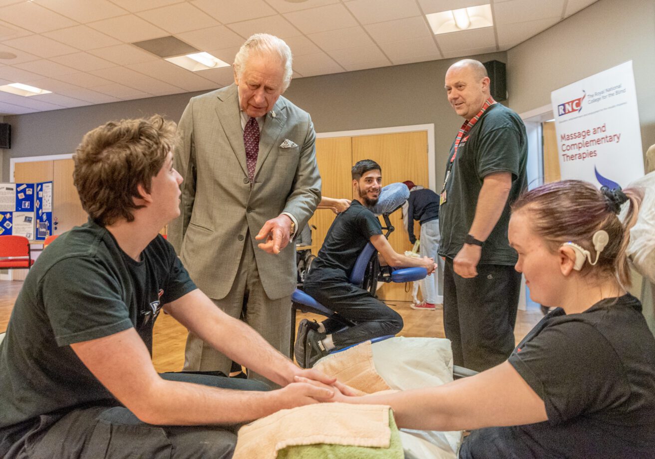 In the foreground two trainee massage students are demonstrating hand massage. King Charles is talking to one of these students, In the background students are demonstrating chair massage as their teacher watches on