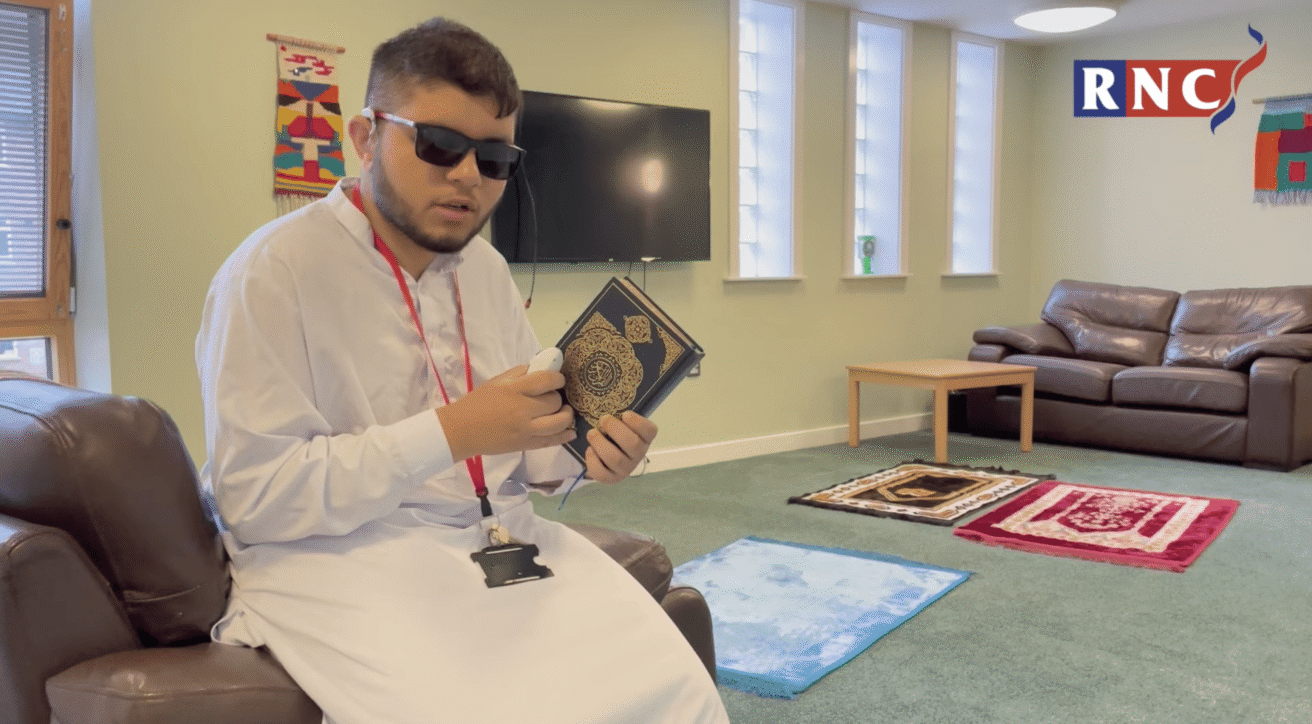 A student wearing traditional dress and holding a copy of the Quran speaks to camera, there are prayer mats on the floor i nthe background
