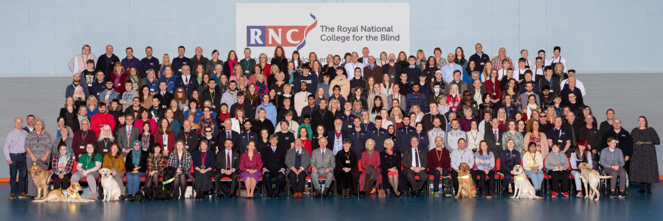 A huge group of staff, students, volunteers, governors and guide dogs sitting on bleacher seating with the King in the centre of the front row