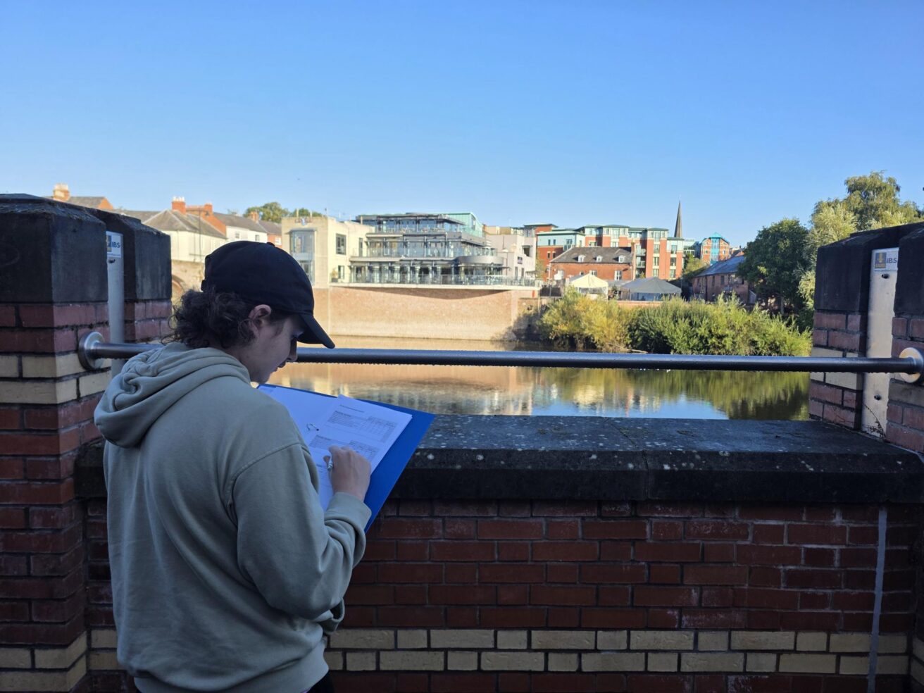 A person wearing a hoodie and cap writing on a clipboard while standing on a brick bridge overlooking a river and town buildings.