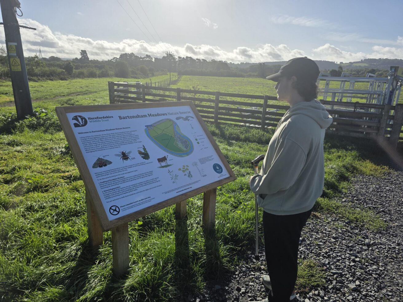 A person standing outdoors next to a wooden information board for the Bartonsham Meadows Nature Reserve.