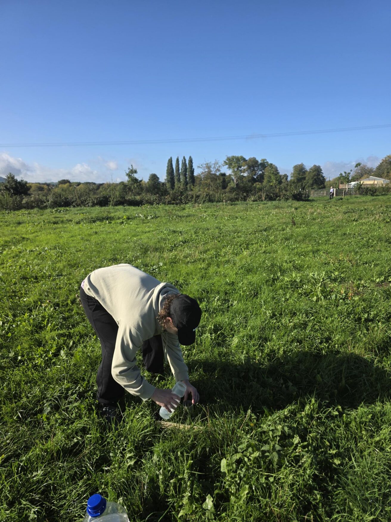 A person bending down in a grassy field pouring water from a bottle onto the ground.
