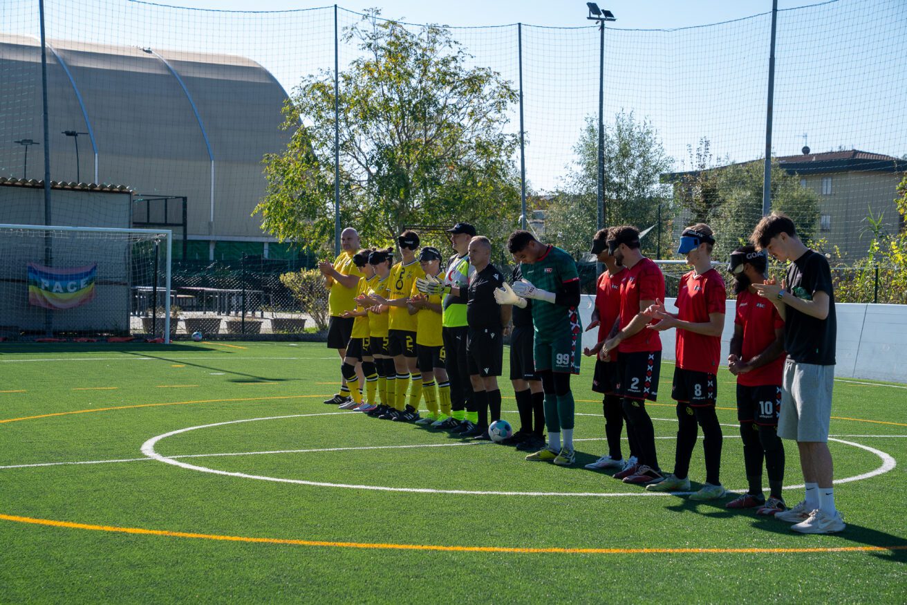 An outdoor, wide shot of two blind football teams standing in a line along the center circle of a green artificial turf pitch on a sunny day. One team on the left wears yellow jerseys and black shorts, while the team on the right wears red jerseys and dark shorts, with eye shades on the outfield players. Goalkeepers, referees, and coaches stand alongside them. All participants are clapping in sportsmanship.