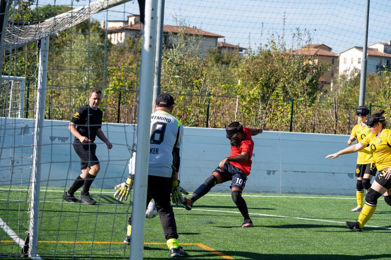 An action shot taken from behind the goalposts on an outdoor artificial turf pitch during a blind football match. In the centre, a blindfolded player wearing a red jersey (number 10) and dark shorts winds up to strike the ball toward the net. A goalkeeper wearing a grey and black Dortmund kit with number 18 stands in front of the goal, watching the play unfold. On the right, two opposing players in yellow Borussia Dortmund jerseys observe the attack, while a referee in black watches from the left background.